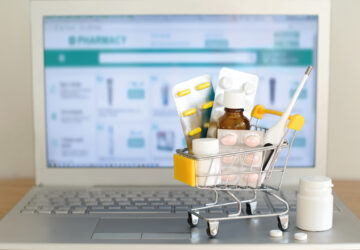 Shopping Cart Toy With Medicaments In Front Of Laptop Screen With Pharmacy Web Site On It. Pills, Blister Packs, Medical Bottles, Thermometer Set. Health Care And Internet Shopping.